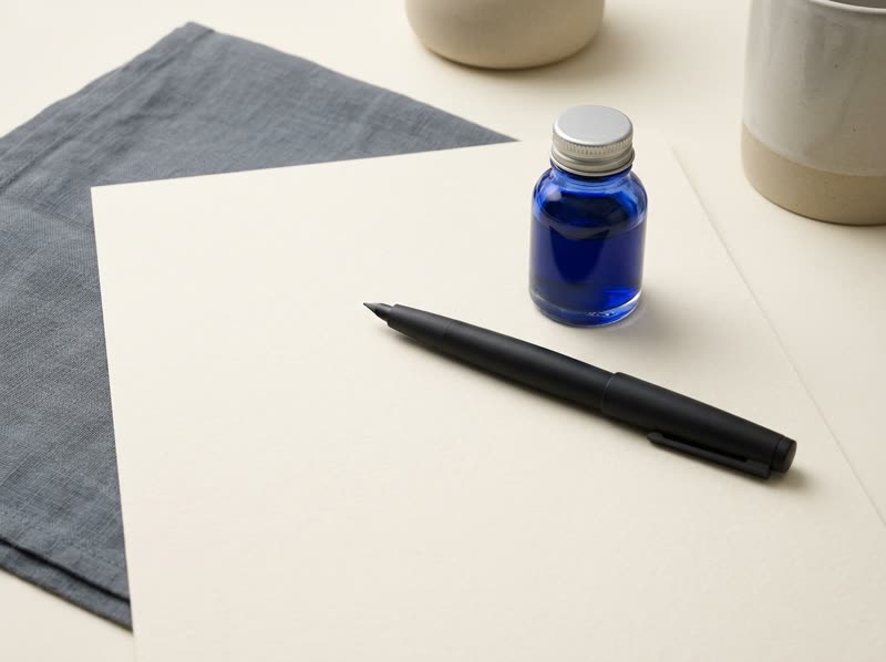 A pen resting on blank paper beside a small bottle of blue ink.
